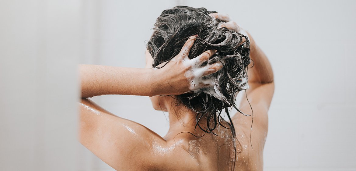 Frau mit dem Rücken zur Kamera wäscht sich in der Dusche mit Shampoo die Haare.